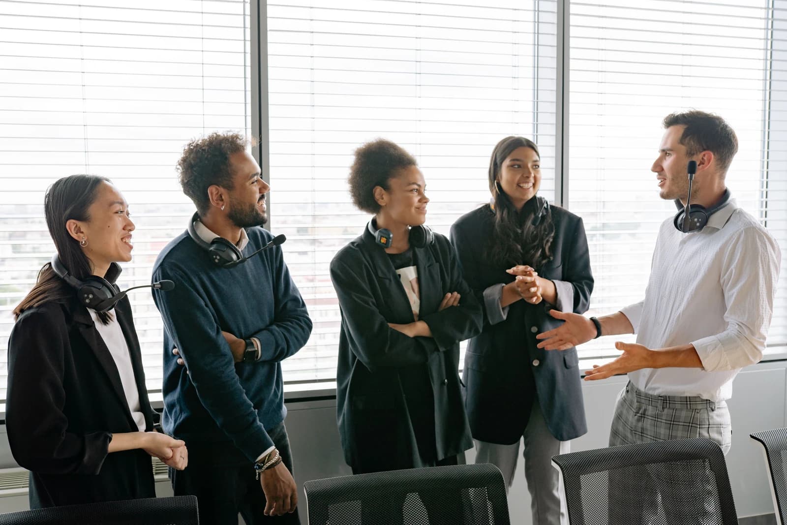 Five colleagues wearing headsets listen to a coworker speaking in an office.