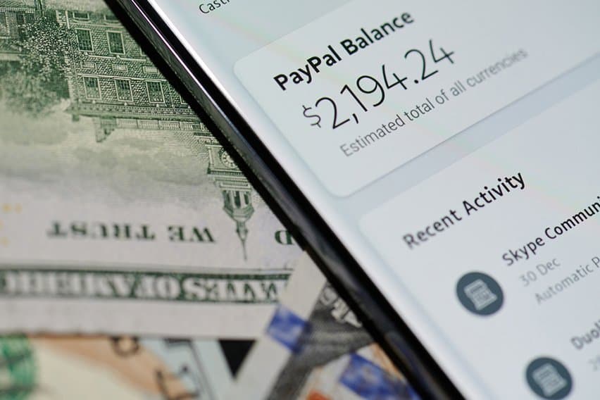 A smartphone showing a PayPal balance of $2,194.24 placed over US dollar bills.
